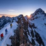 Trois alpinistes progressent sur une crête neigeuse escarpée au lever du soleil avec la Barre des Écrins en arrière-plan.