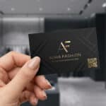 Comment créer une carte visite mode mémorable en 2026 ? Main tenant une carte de visite noire élégante avec logo doré "AF" et QR code dans une boutique de mode moderne.
