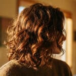 Femme aux cheveux bouclés châtain cuivré illuminés par la lumière dorée du soleil.
